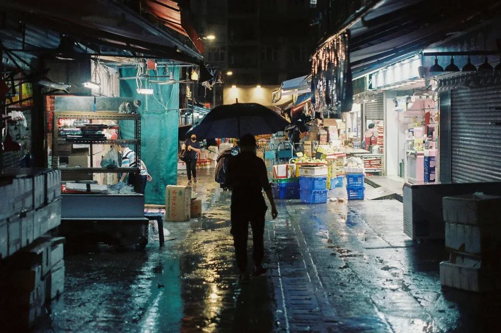 A person carrying an umbrella walks through a dimly lit Singapore wet market at night, with rain-slick floors reflecting the lights, open food stalls, produce crates, and vendors along both sides.