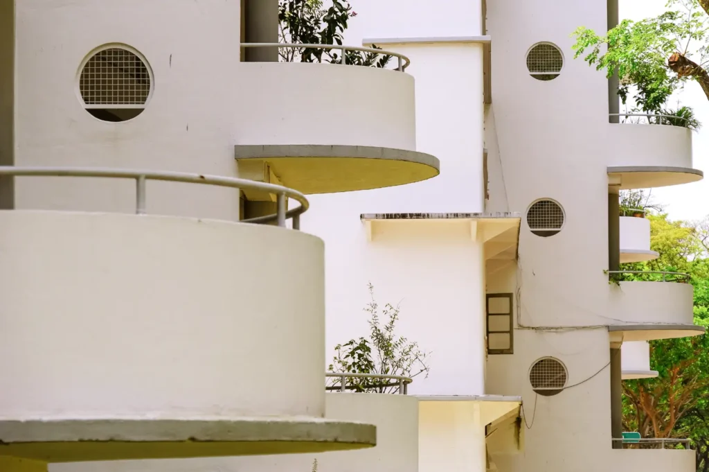 White multi-story apartment facade with curved balconies, railings, and small round ventilation grilles set into the walls, surrounded by green trees and plants.