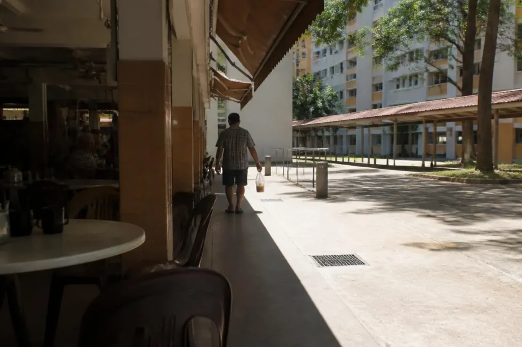 A café-lined covered walkway in Singapore with harsh midday sunlight, bright concrete pavement, deep shadows, tropical trees, and a person walking in the distance.