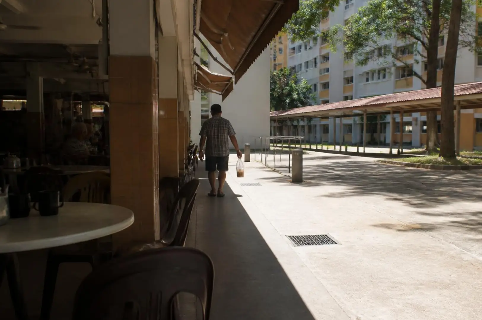 A café-lined covered walkway in Singapore with harsh midday sunlight, bright concrete pavement, deep shadows, tropical trees, and a person walking in the distance.