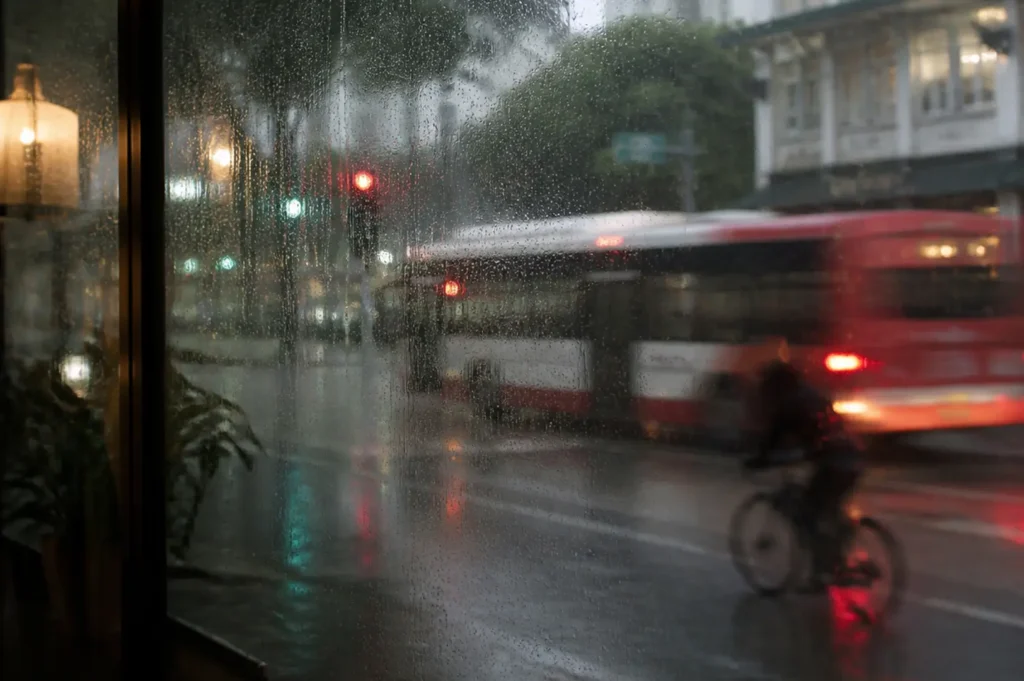 A rainy Singapore street viewed through a café window, with a red bus passing through the wet roadway and a cyclist riding alongside it. Reflections from traffic lights shimmer on the pavement, while trees and high-rise buildings appear softly blurred in the background.