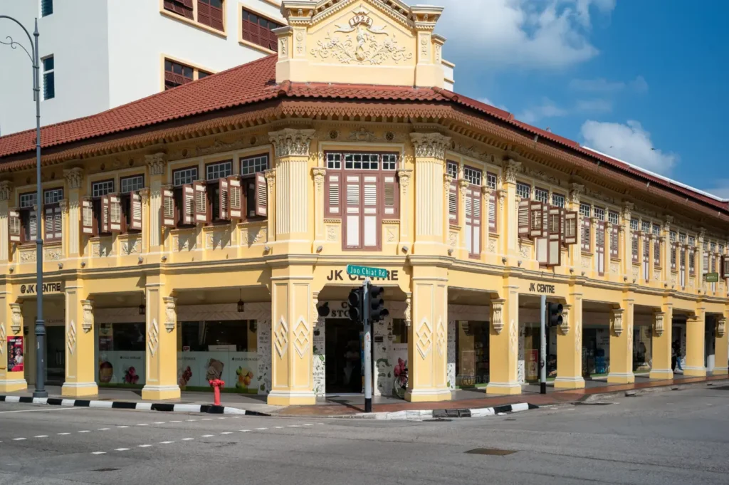 Historic yellow building with arched windows and ornate balcony details along a street, with a few people walking and a blue sky with clouds overhead.