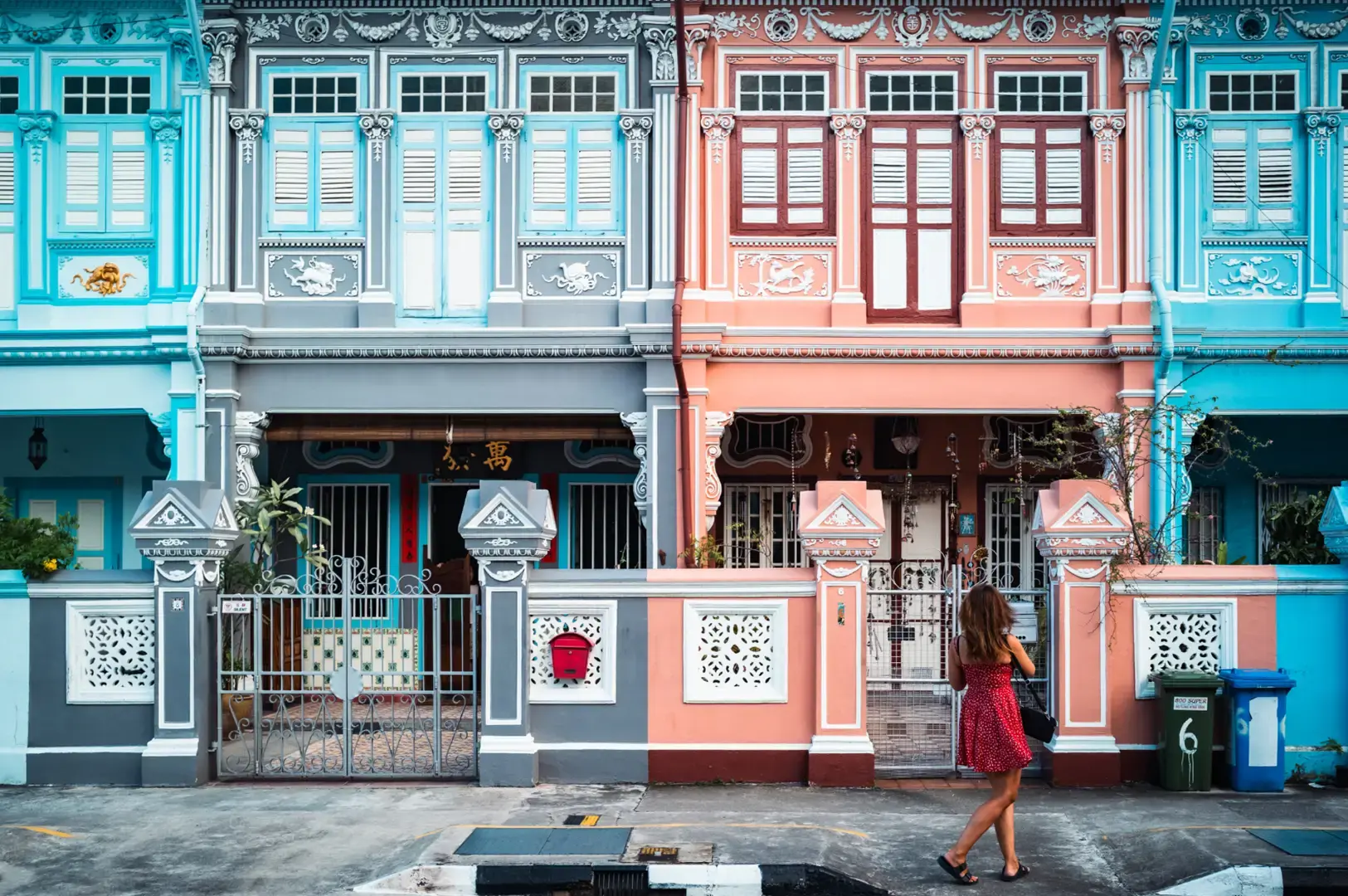 Colorful Joo Chiat shophouses with ornate façades, blue and pink façades, tiled entrances, and a woman walking along the street.