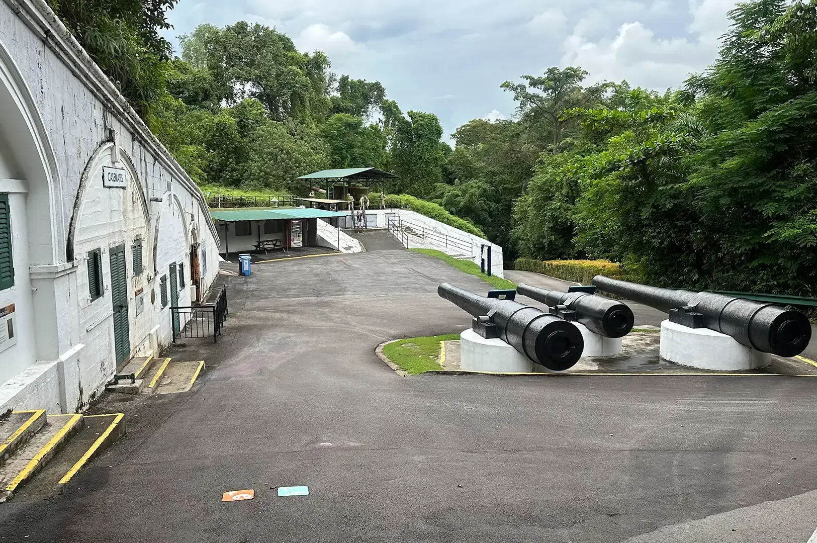 Fort Siloso grounds with three historic cannons outside a white fort building, surrounded by dense greenery.
