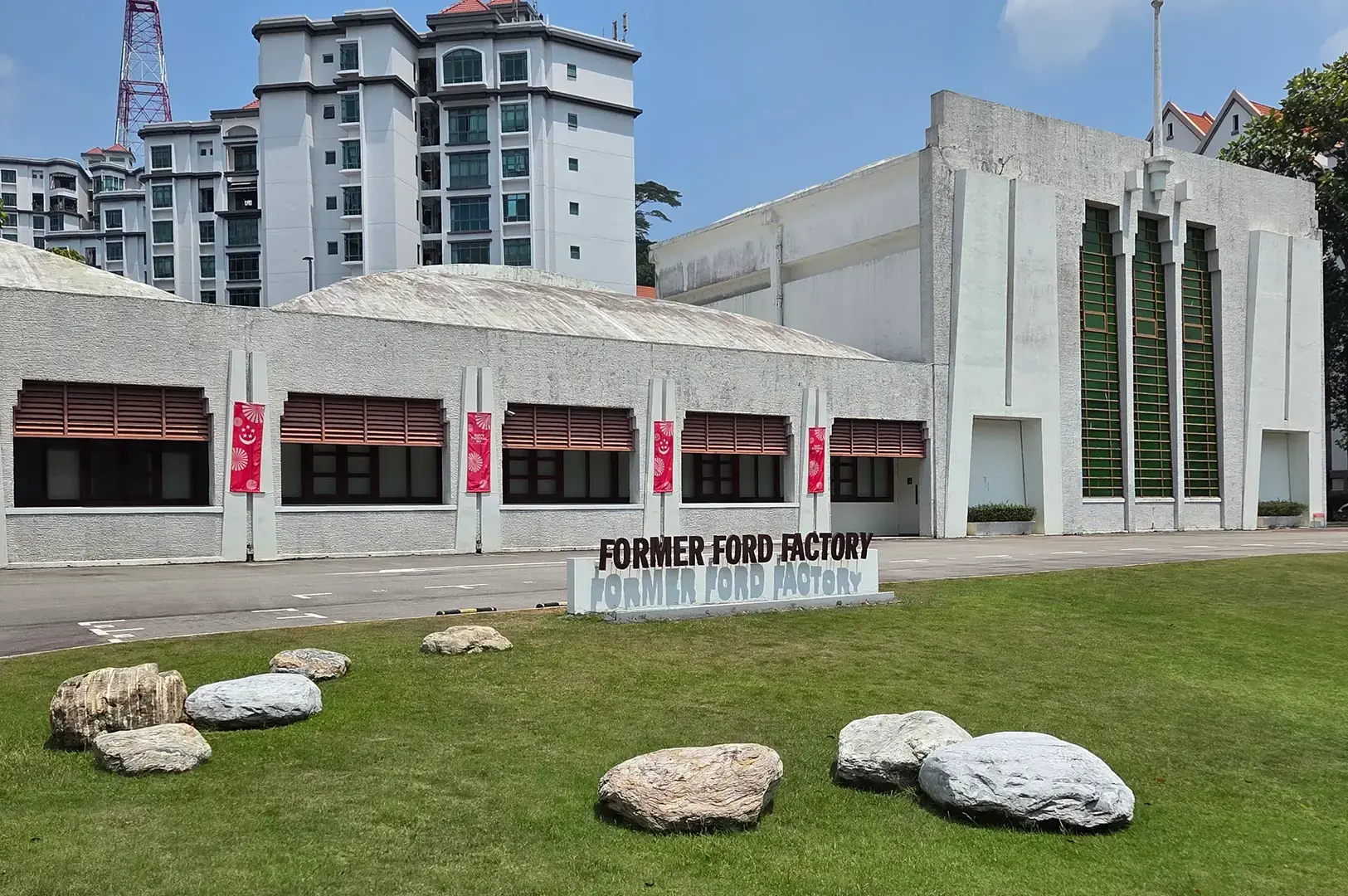 Former Ford Factory building with white façades, red banners, a lawn scattered with stones, and the “Former Ford Factory” sign.