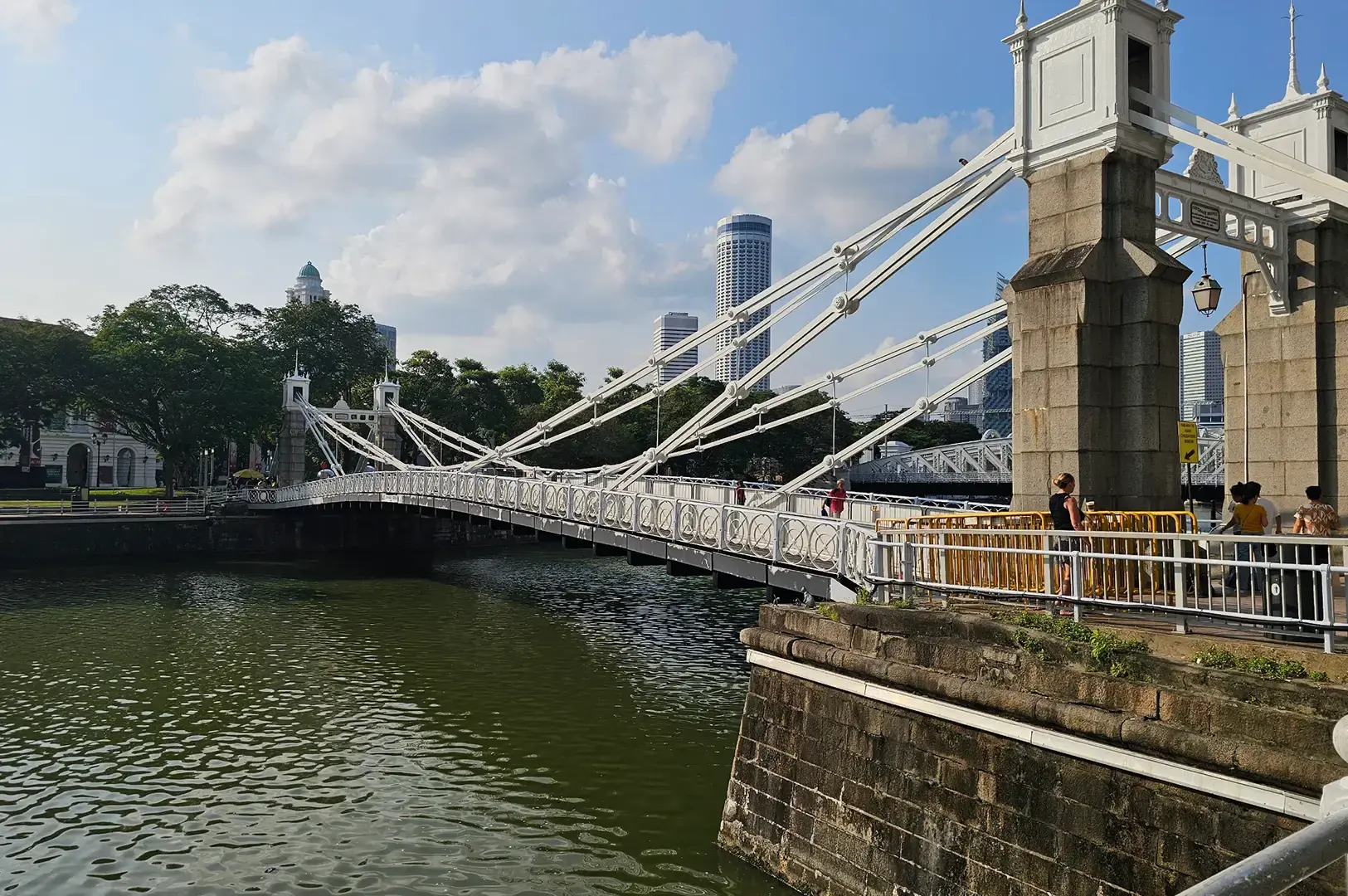 Cavenagh Bridge spanning the Singapore River, with historic suspension towers, pedestrians, riverside walkways, and the city skyline in the background.