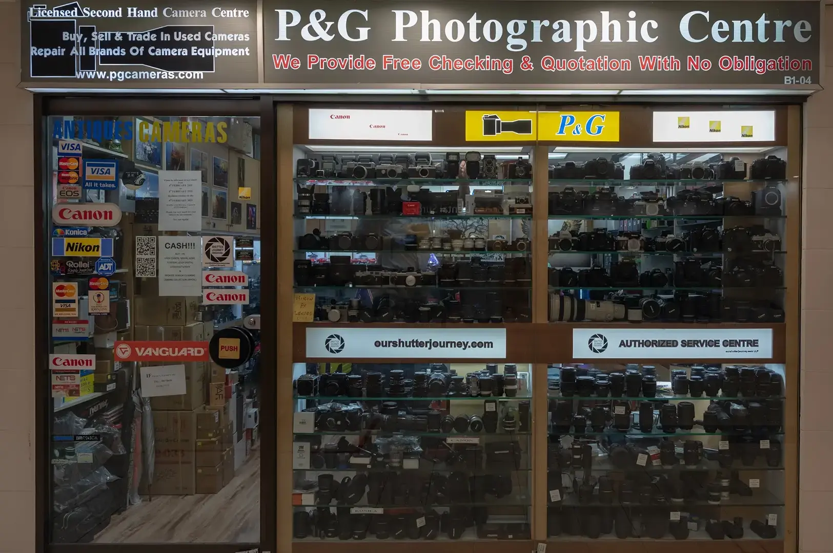 P&G Photographic Centre storefront displaying new and second-hand cameras, lenses, accessories, and repair service signs.